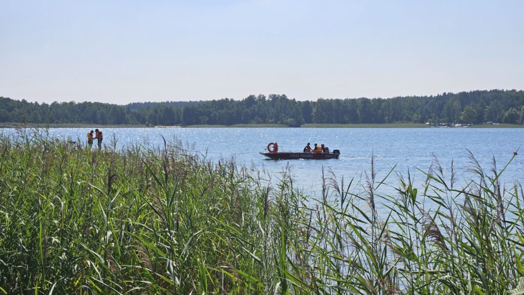 Dwóch mężczyzn w kamizelkach odblaskowych rozmawia nad brzegiem jeziora, obok płynie łódź straży pożarnej.