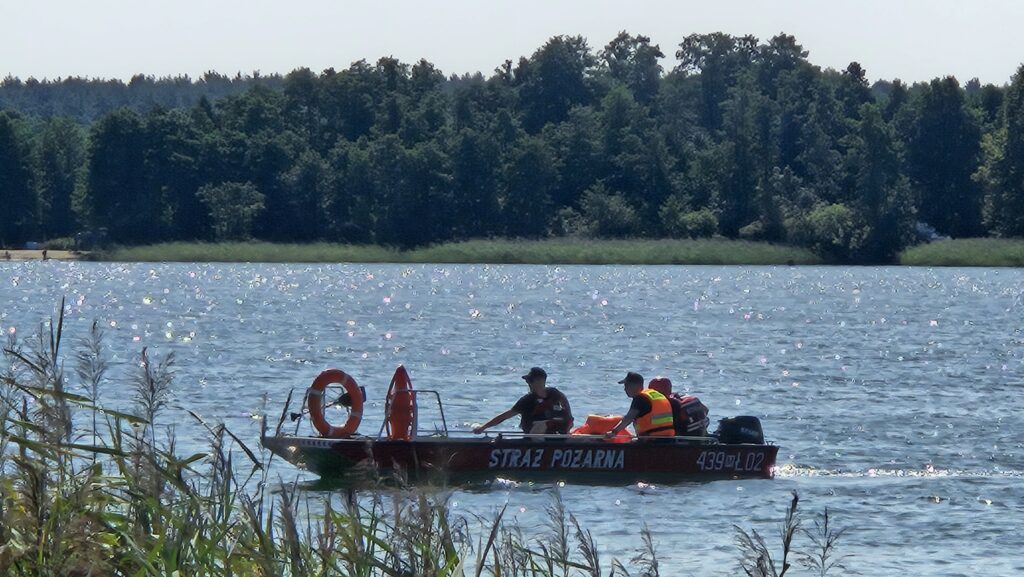 Strażacy na łodzi motorowej patrolują wodę w słoneczny dzień, w tle zalesiony brzeg.