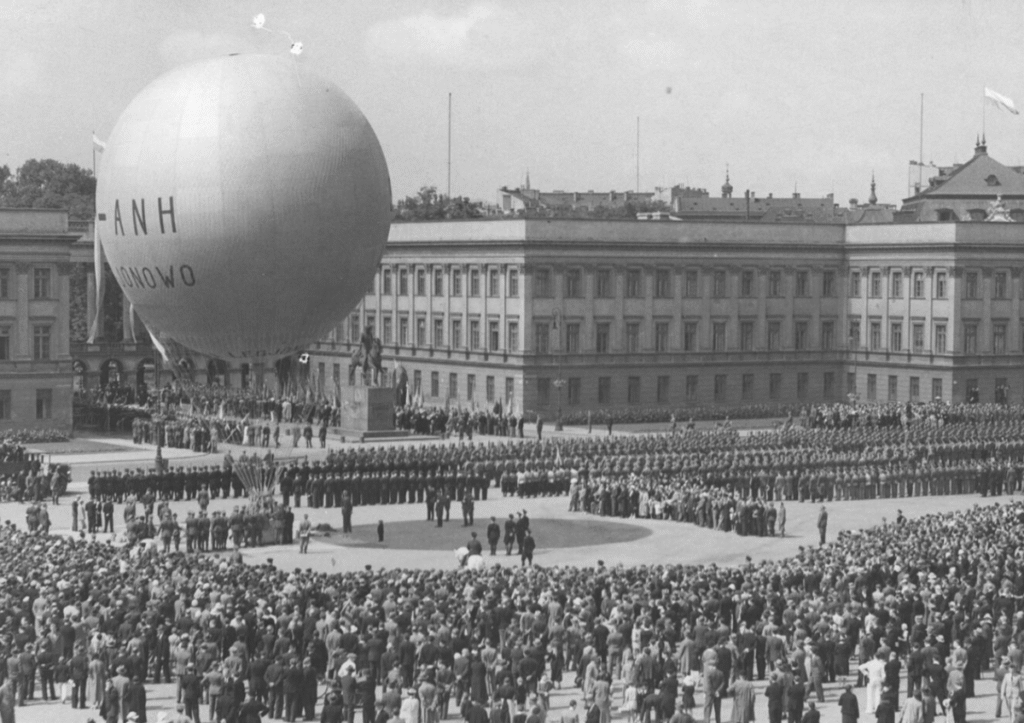 Tłum tłumu obserwuje wydarzenie z balonem unoszącym się nad placem, w tle monumentalny budynek.