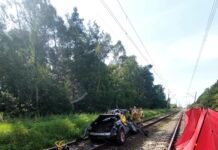 Zniszczony samochód stoi na torach kolejowych, obok grupy strażaków zajmujących się akcją ratunkową. W tle widoczna zieleń drzew.