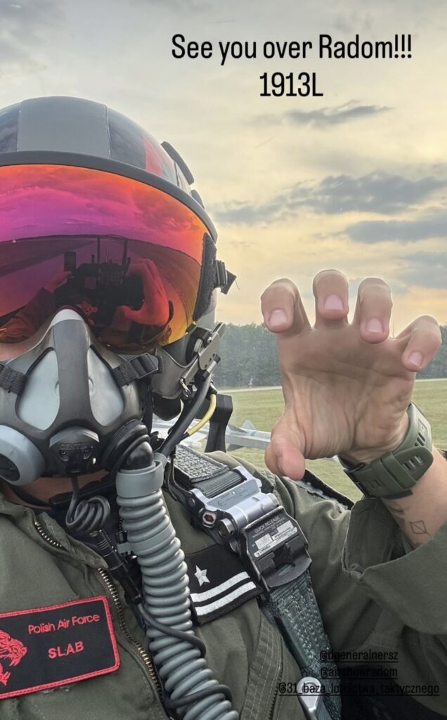 Pilot w hełmie i masce tlenowej wykonuje gest w stronę kamery na tle nieba z chmurami.