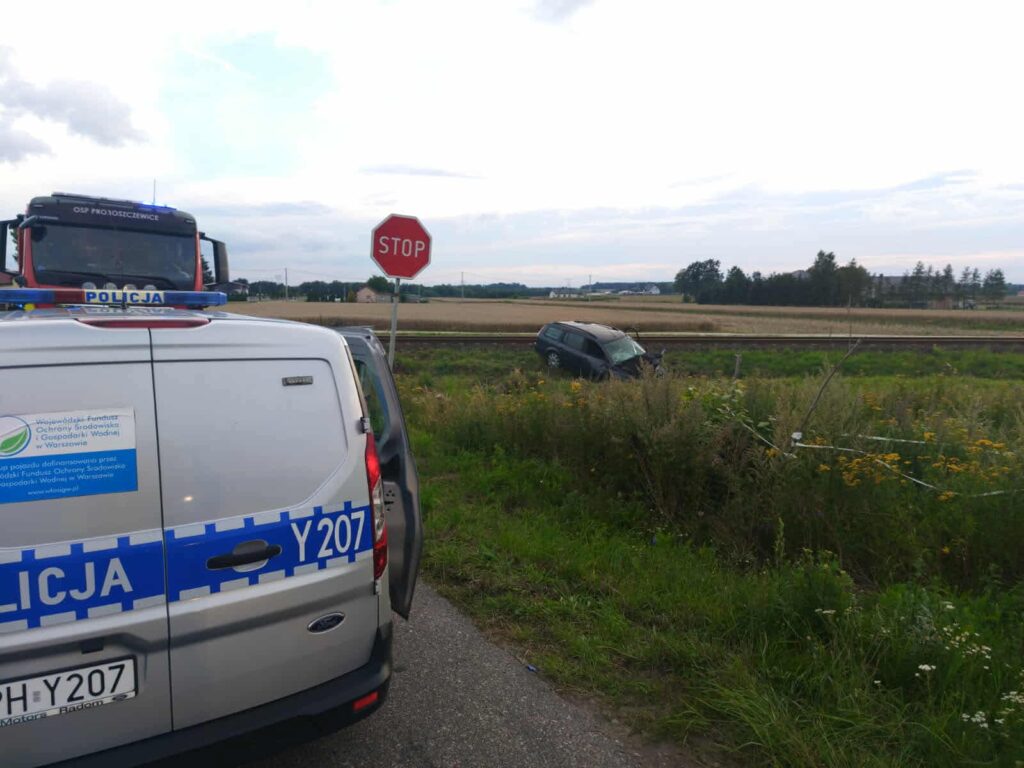 Policyjny radiowóz z widocznym emblematem stoi na drodze obok przewróconego samochodu w polu. Na tle widoczne znaki drogowe.