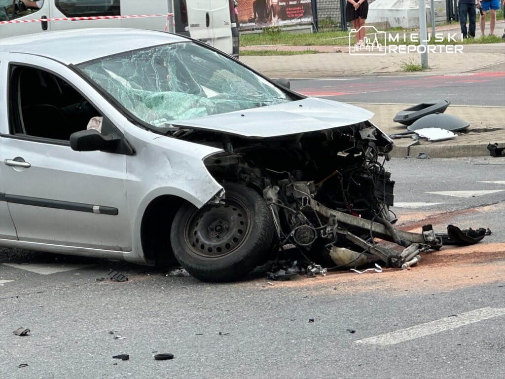 Uszkodzony samochód osobowy z rozbitą przednią częścią stoi na jezdni, wokół leżą fragmenty pojazdu.