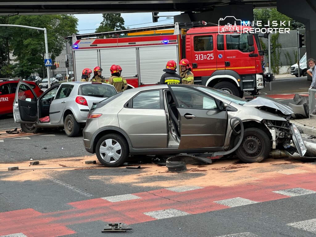 Dwa uszkodzone samochody na skrzyżowaniu, obok strażacy w hełmach rozmawiający przy wozie strażackim.