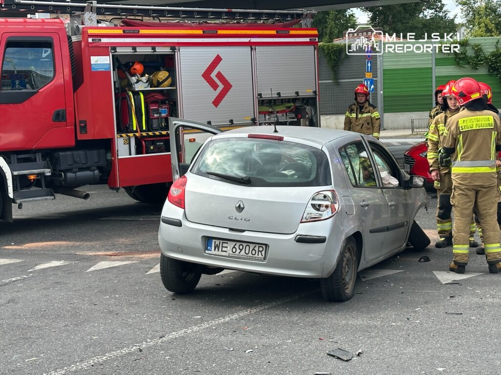 Strażacy w hełmach rozmawiają obok uszkodzonego samochodu osobowego na skrzyżowaniu.