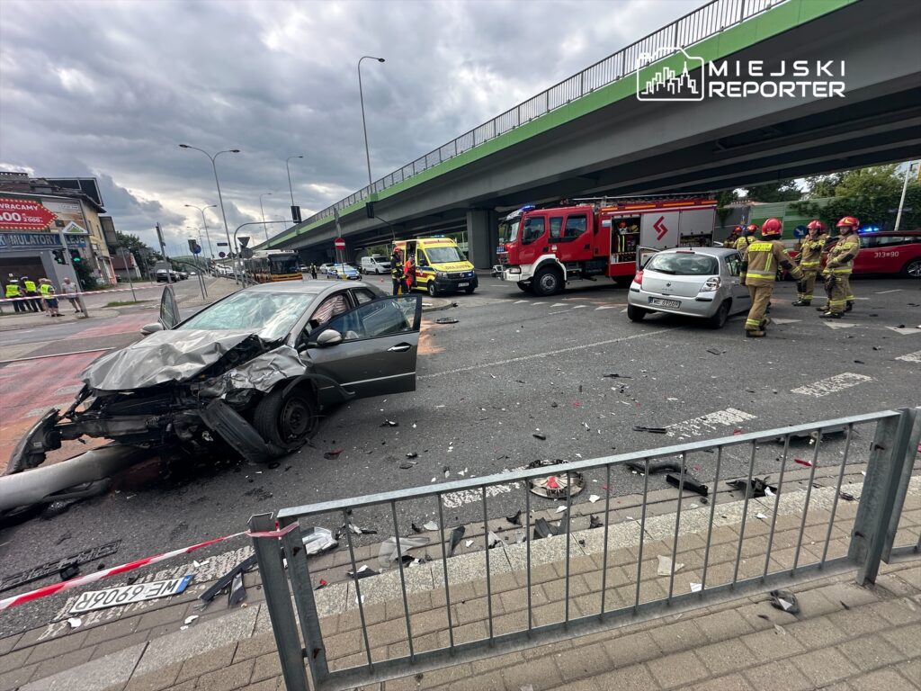 Zniszczony samochód osobowy stoi na drodze, obok strażacy oraz wóz strażacki, w tle ruch uliczny.