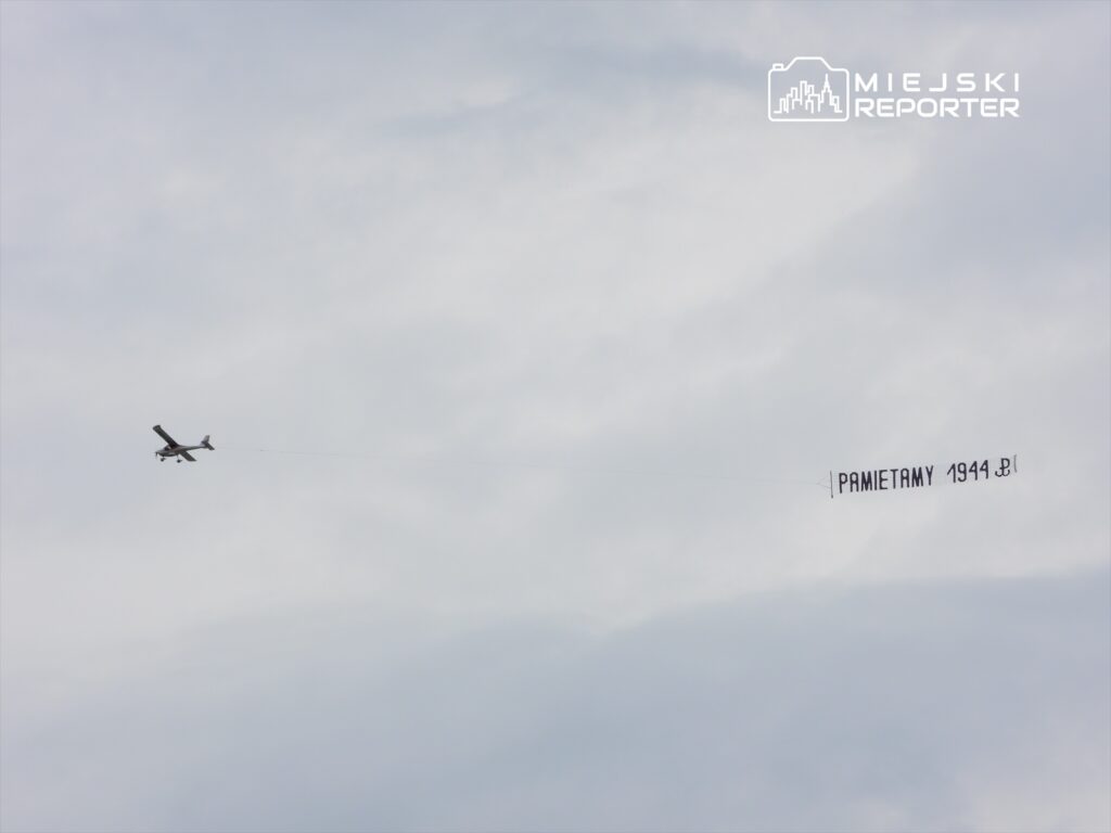 Samolot przeciągający transparent z napisem "PAMIĘTAMY 1944" na tle pochmurnego nieba.