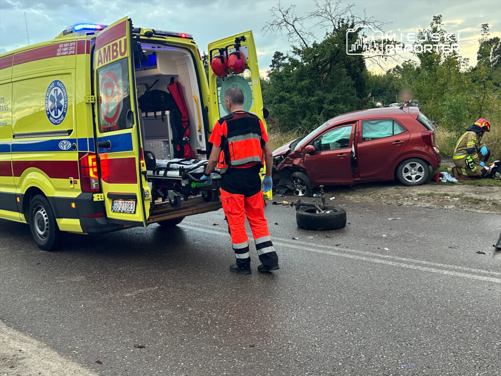 Ratownicy medyczni przygotowują nosze przy otwartej tylnej części karetki obok uszkodzonego czerwonego samochodu osobowego.