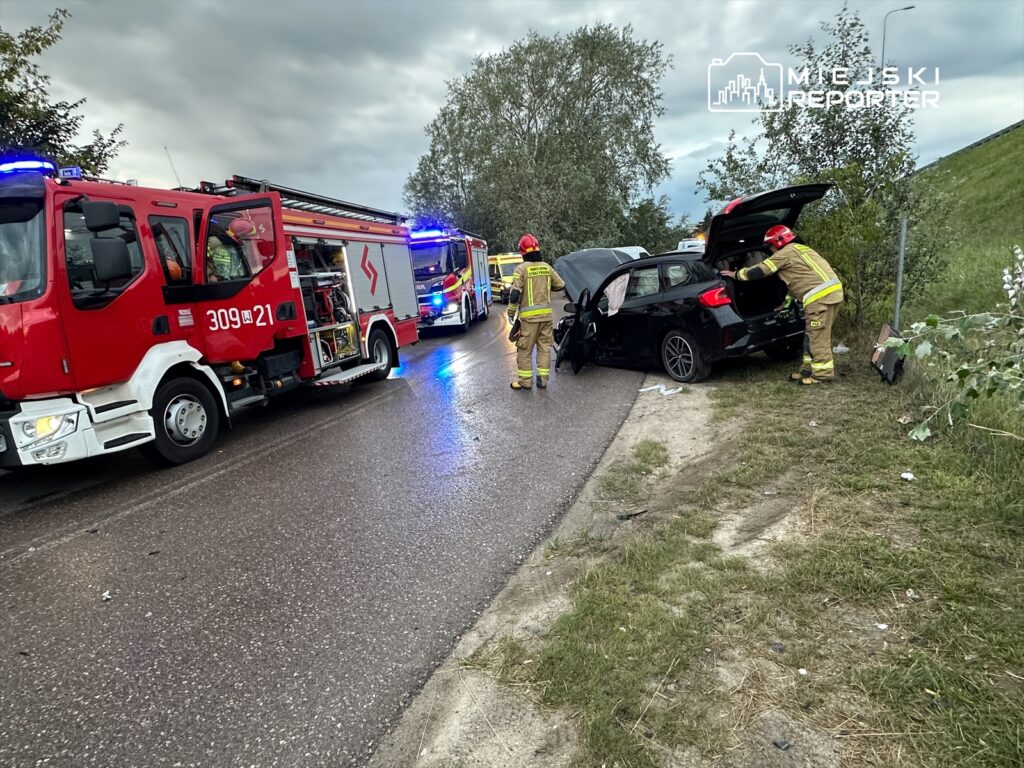 Strażacy w mundurach ratunkowych sprawdzają uszkodzony samochód osobowy przy drodze.