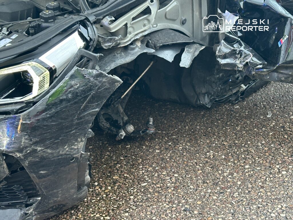 Uszkodzony przód samochodu z wygiętą karoserią i uszkodzoną lampą, leżący na drodze.