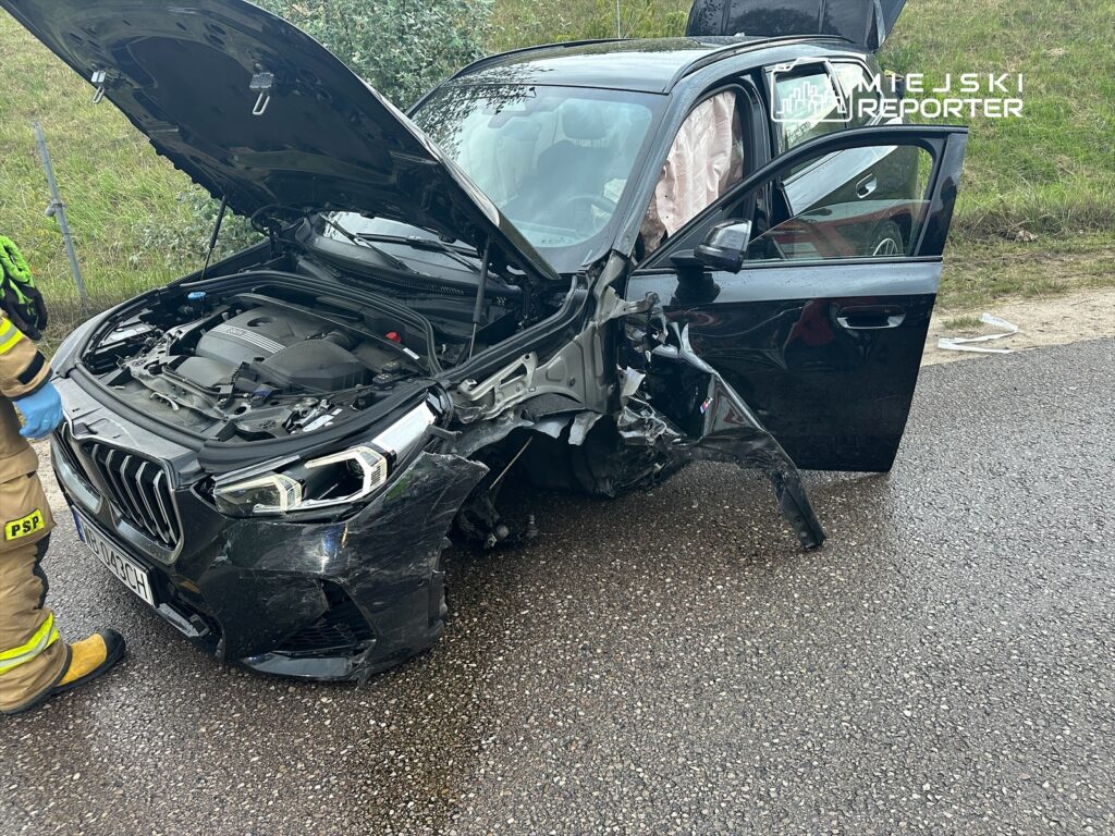 Zniszczony czarny samochód osobowy z otwartą maską, obok strażak w mundurze bada uszkodzenia pojazdu na drodze.
