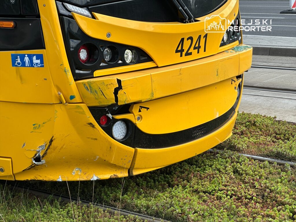 Uszkodzony żółty tramwaj z widocznymi zadrapaniami i wgnieceniami, stojący na torowisku w otoczeniu zieleni.