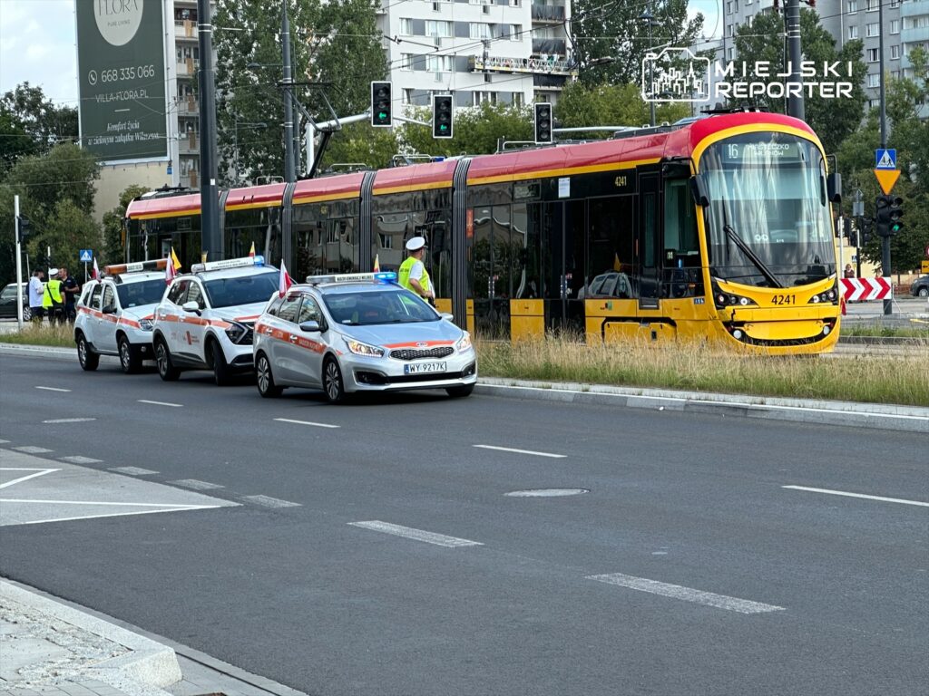 Grupa policjantów kieruje ruchem drogowym obok tramwaju, a za nimi stoją trzy radiowozy z włączonymi sygnałami świetlnymi.