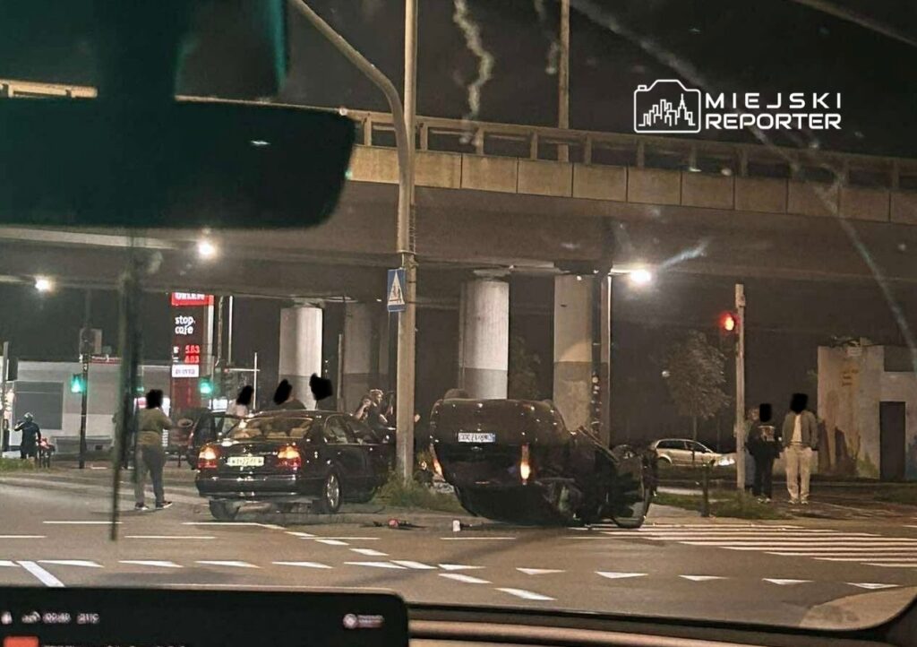 Na skrzyżowaniu ulic, przewrócone auto leży na dachu, a obok grupka osób obserwuje sytuację. W tle widać sygnalizację świetlną.