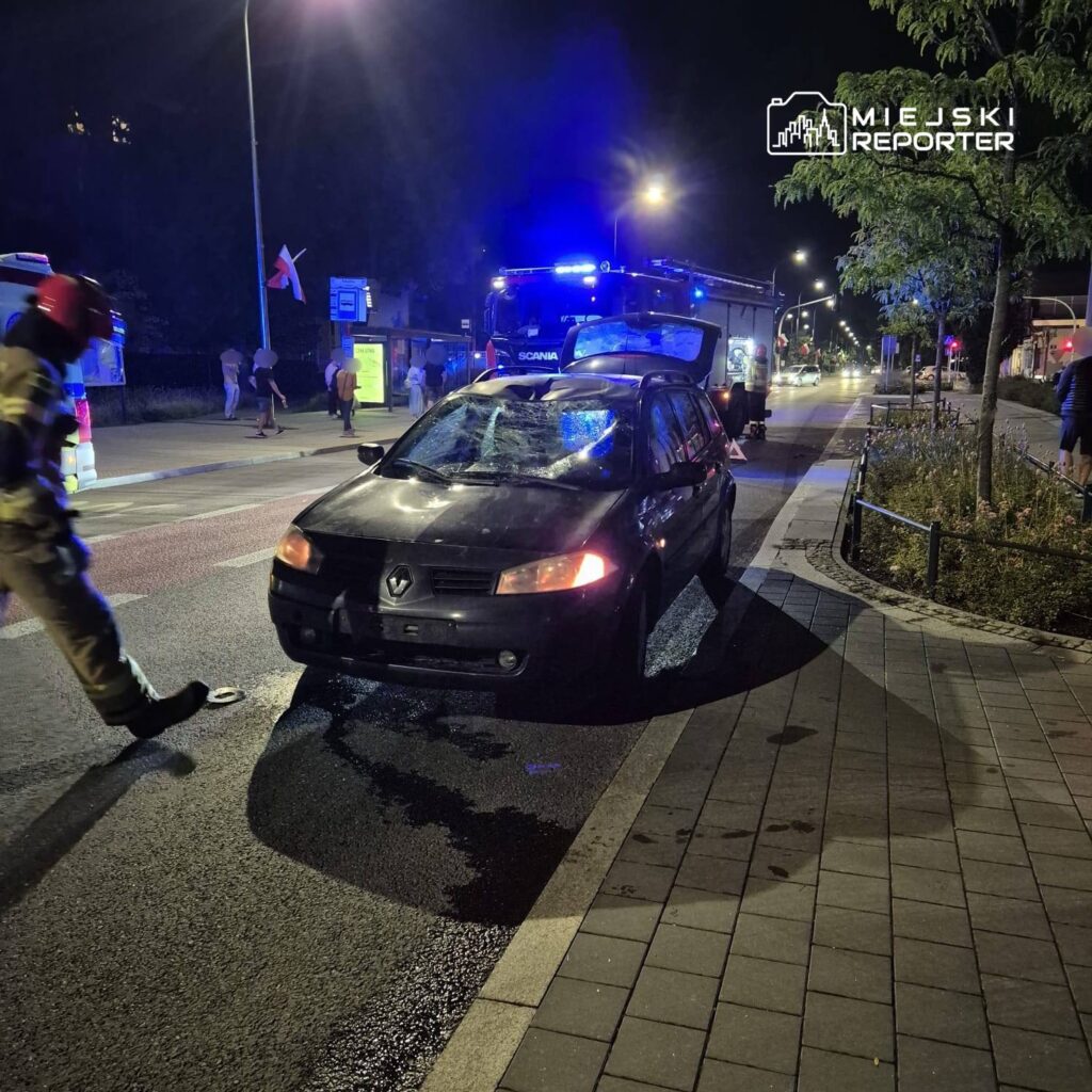 Strażak w hełmie ocenia uszkodzone auto na ulicy w nocy, obok widoczny wóz strażacki z włączonymi sygnalizatorami.