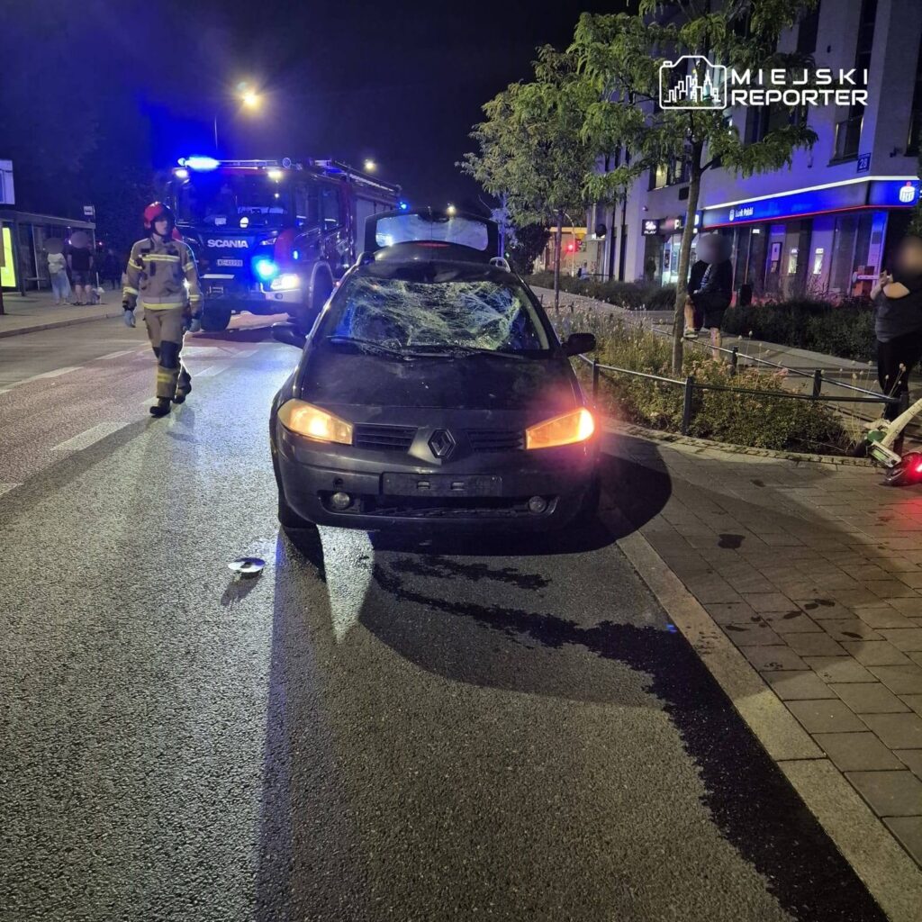 Czarny samochód osobowy z uszkodzoną przednią szybą stoi na ulicy, obok strażacy w mundurach.