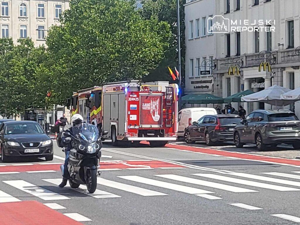 Motocyklista przejeżdża obok wozu strażackiego z włączonymi sygnałami świetlnymi na ulicy z zielonymi drzewami w tle.