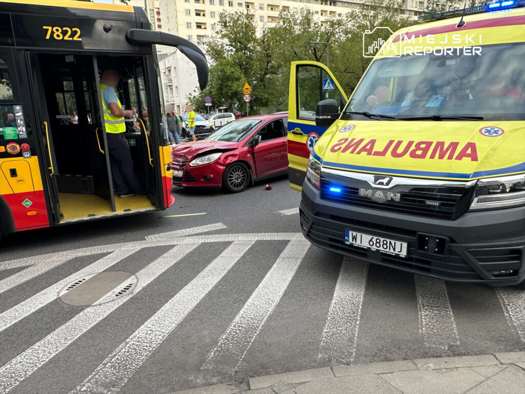 Kierowca autobusu w kamizelce odblaskowej obserwuje wypadek, w którym czerwony samochód osobowy zderzył się z pojazdem.