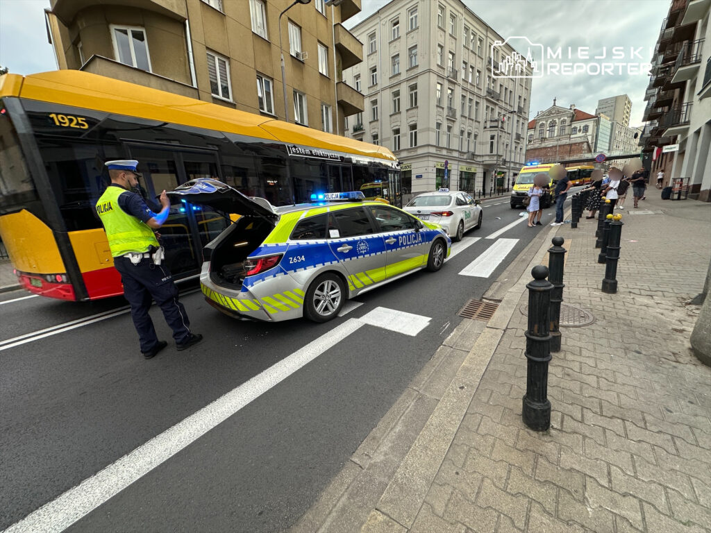 Policjant w kamizelce odblaskowej przeszukuje bagażnik radiowozu na ruchliwej ulicy, obok zatrzymany autobus.