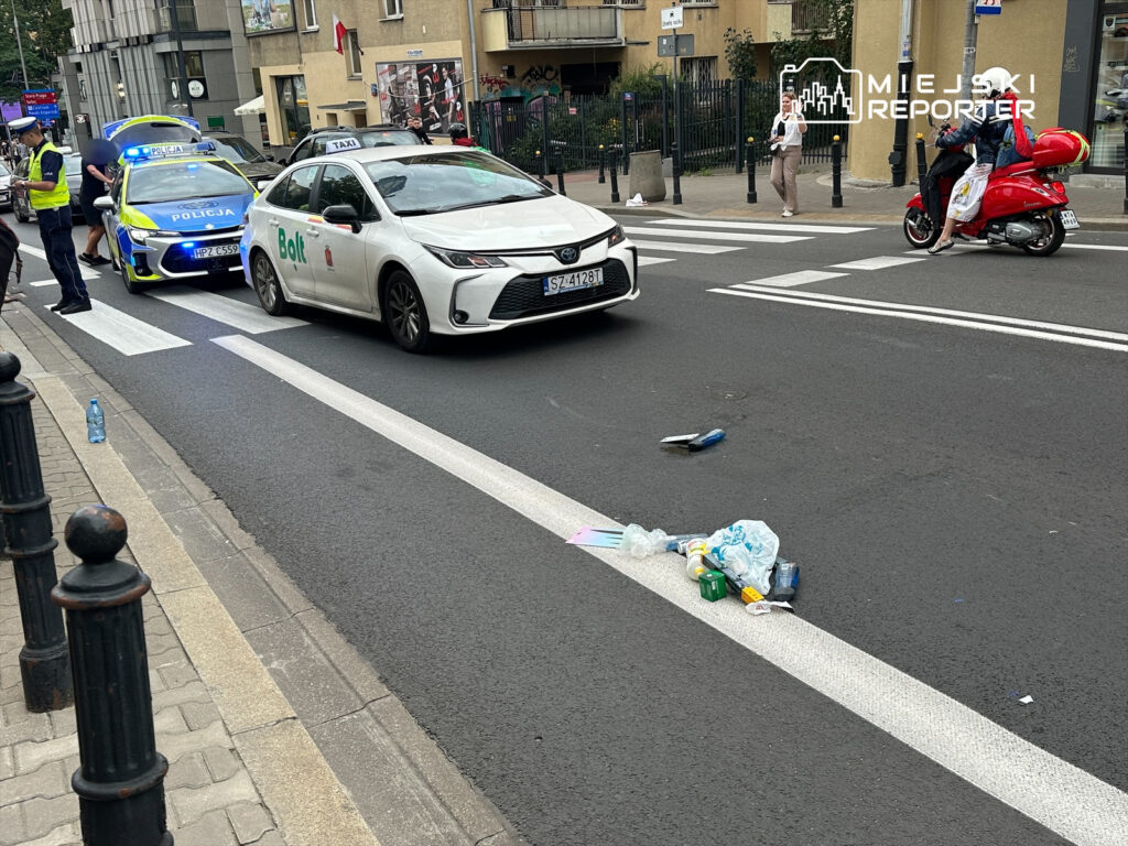Na ulicy zatrzymane są dwa samochody, w tym taksówka i auto policyjne z włączonymi sygnalizatorami. Na jezdni leży porzucony zbiór odpadów.