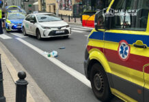 Na ulicy leżą porozrzucane odpady, obok zatrzymane samochody, w tym taksówka i radiowóz policji, a w tle widać ambulans.