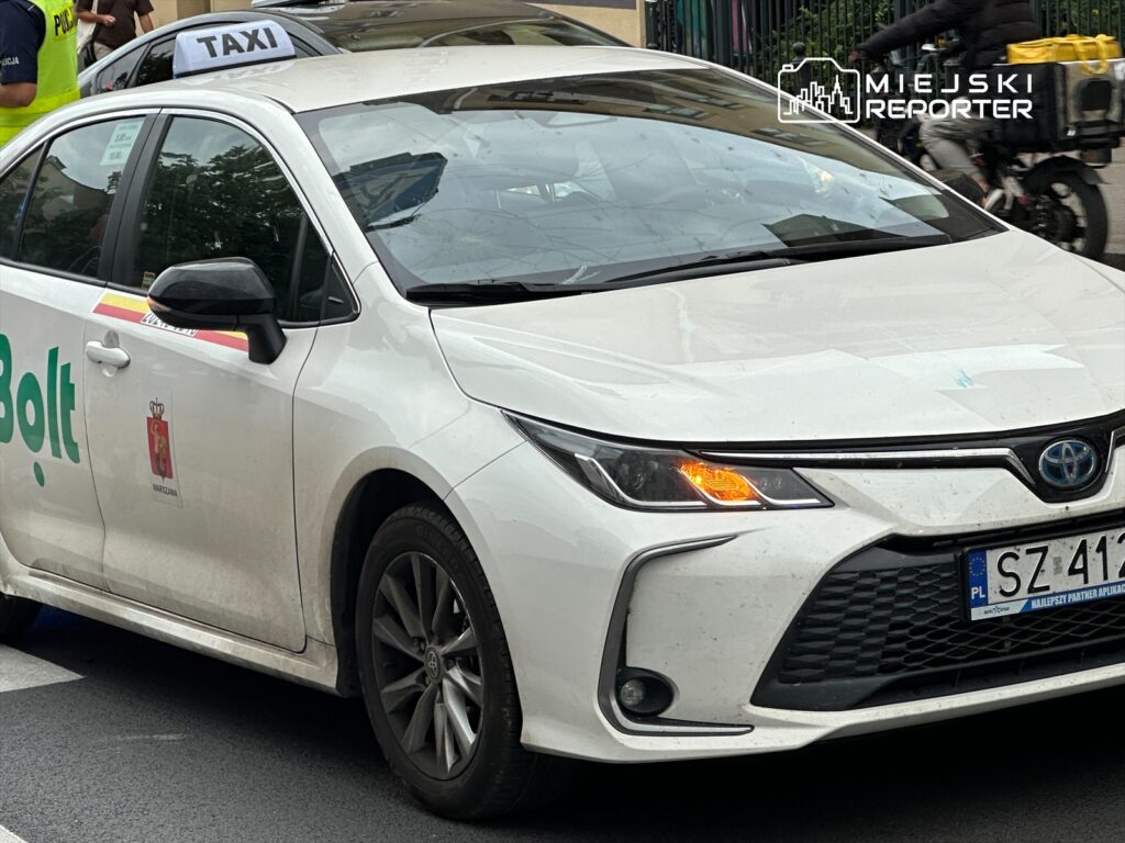 Biały samochód osobowy marki Toyota z logo Bolt, zaparkowany na ulicy, obok policjanta i przechodniów.