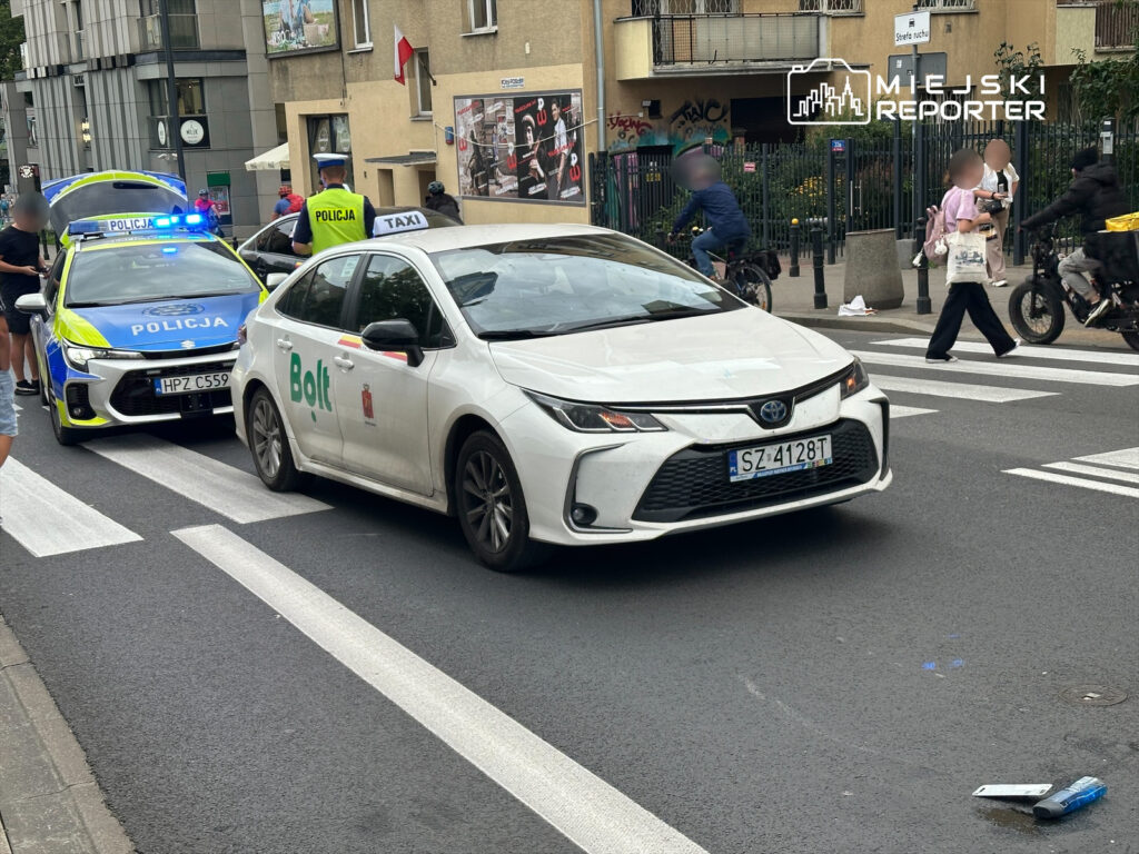 Policyjny radiowóz z włączonymi sygnałami zatrzymuje taksówkę Bolt na ulicy, obok przechodniów i rowerzystów.