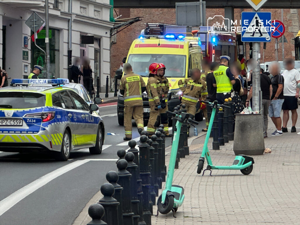Grupa strażaków i policjantów interweniuje na ulicy, obok zaparkowanej karetki pogotowia. Na chodniku stoją elektryczne hulajnogi.