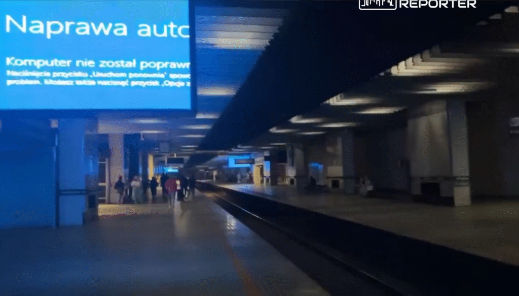 Pasażerowie czekają na peronie w nowoczesnym dworcu kolejowym, a nad nimi wyświetla się komunikat: "Naprwa auto. Komputer nie został poprawiony."