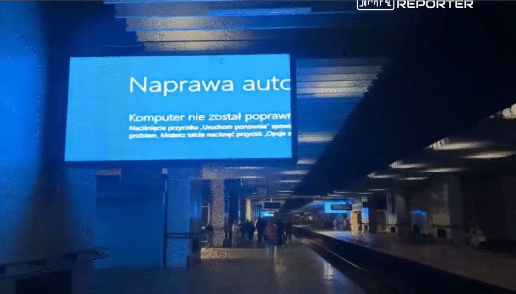 Ekran informacyjny w stacji kolejowej wyświetlający komunikat o błędzie komputera. W tle widać przechodzących pasażerów.