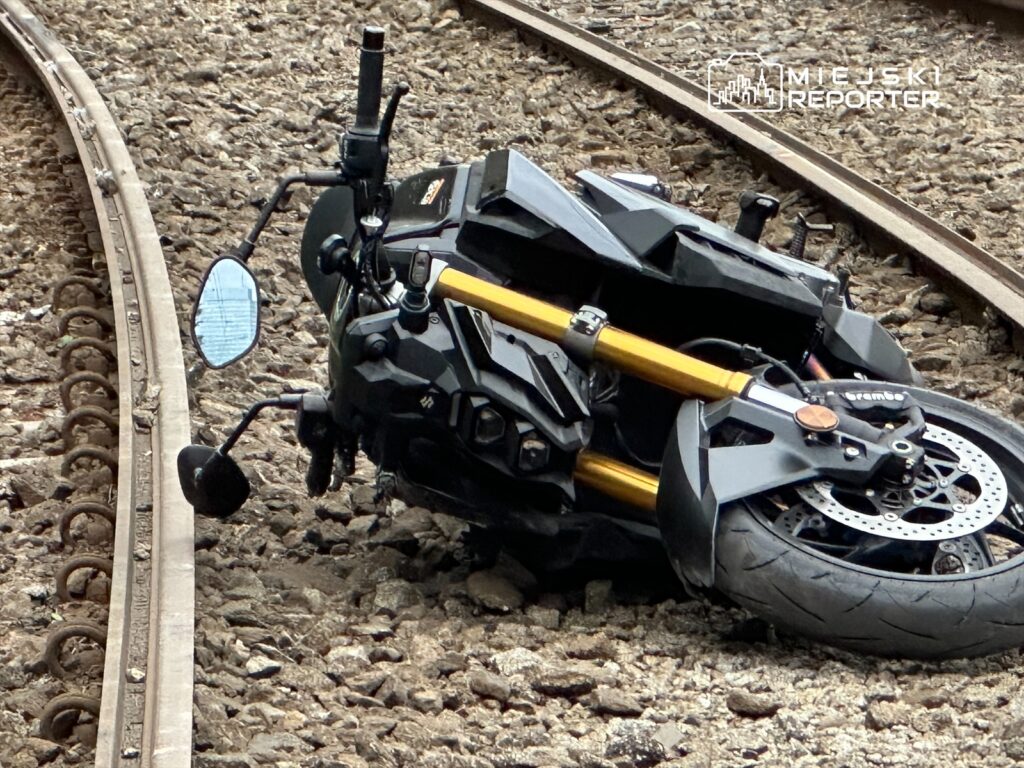 Motocykl leży na torach kolejowych, z uszkodzoną przednią częścią i odwróconym lusterkiem.
