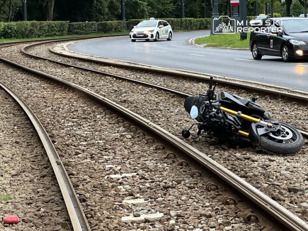 Motocykl leży na torach tramwajowych, obok ruchliwej ulicy, w otoczeniu zieleni i samochodów.