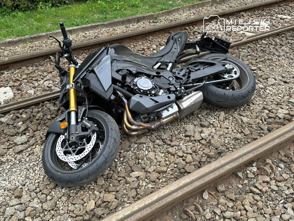 Motocykl leżący na torach kolejowych, z uszkodzoną przednią częścią i oponami. W tle widoczne kamienie i zieleń.