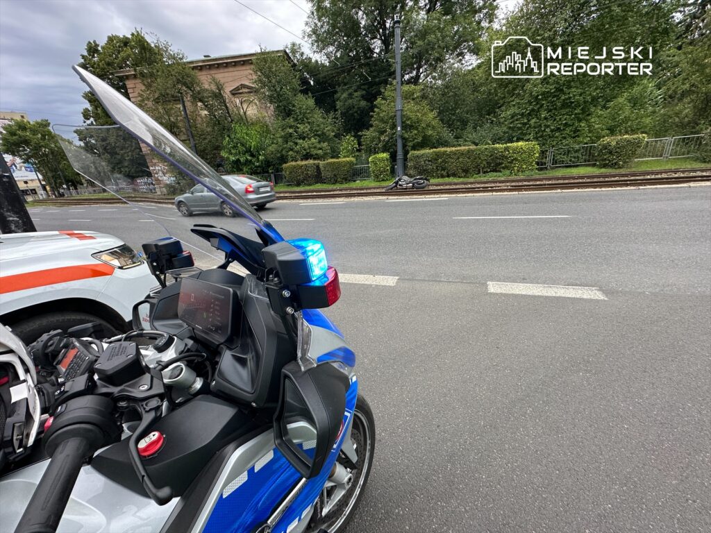 Motocykl policyjny z niebieskim sygnałem świetlnym zaparkowany na ulicy obok radiowozu, w tle zieleń i budynki.