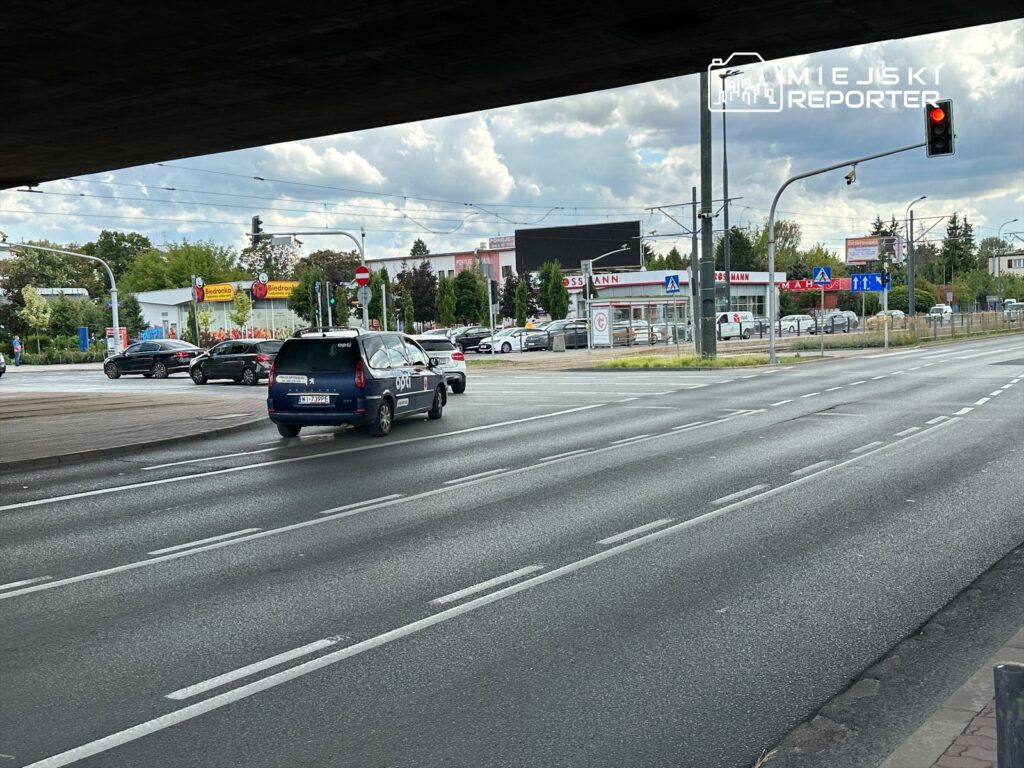 Samochód osobowy skręcający w lewo na skrzyżowaniu, obok widoczne znaki drogowe i sklepy w tle.