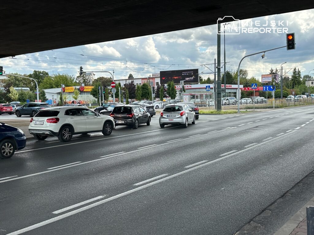 Ruch samochodowy na skrzyżowaniu z sygnalizacją świetlną, w tle widoczne reklamy i budynki handlowe.