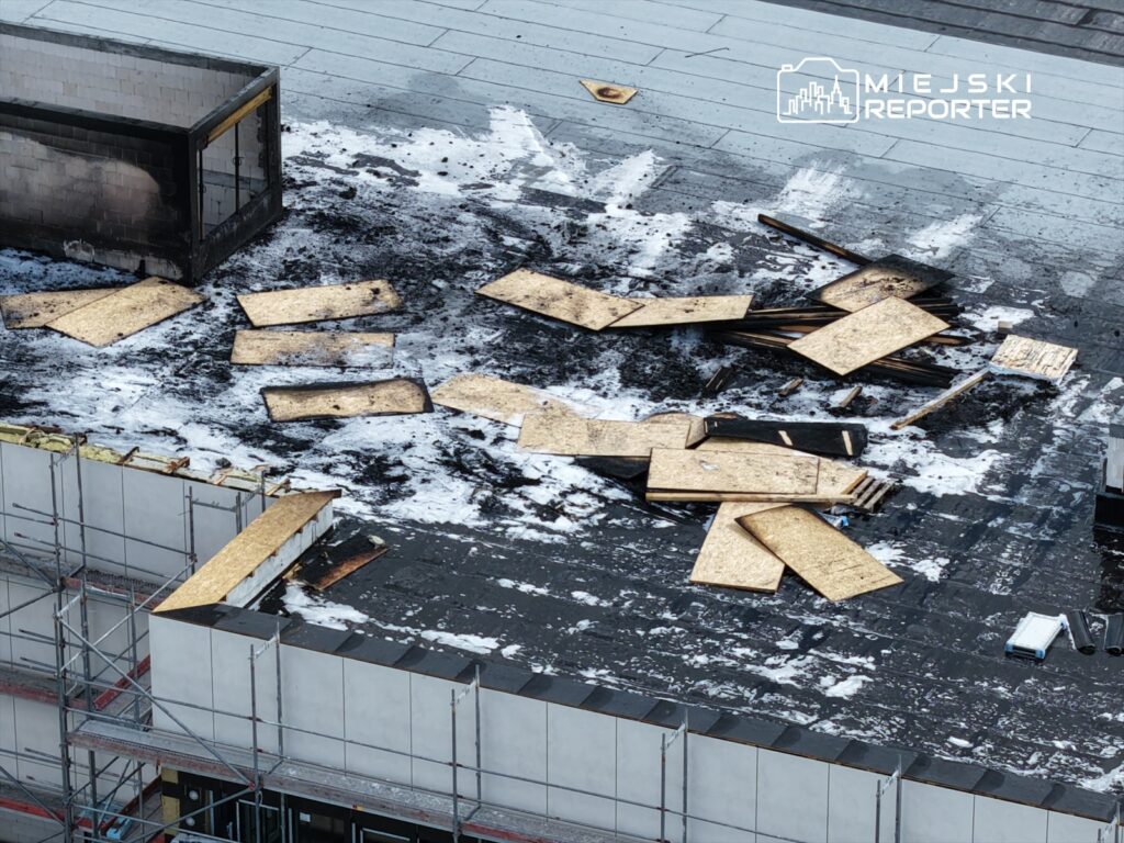 Na dachu budynku leżą spalone płyty OSB oraz czarne zacieki, otoczone materiałami budowlanymi.