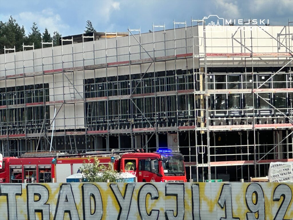 Nowoczesny budynek w trakcie budowy, otoczony rusztowaniami, z zaparkowanymi wozami strażackimi w tle.