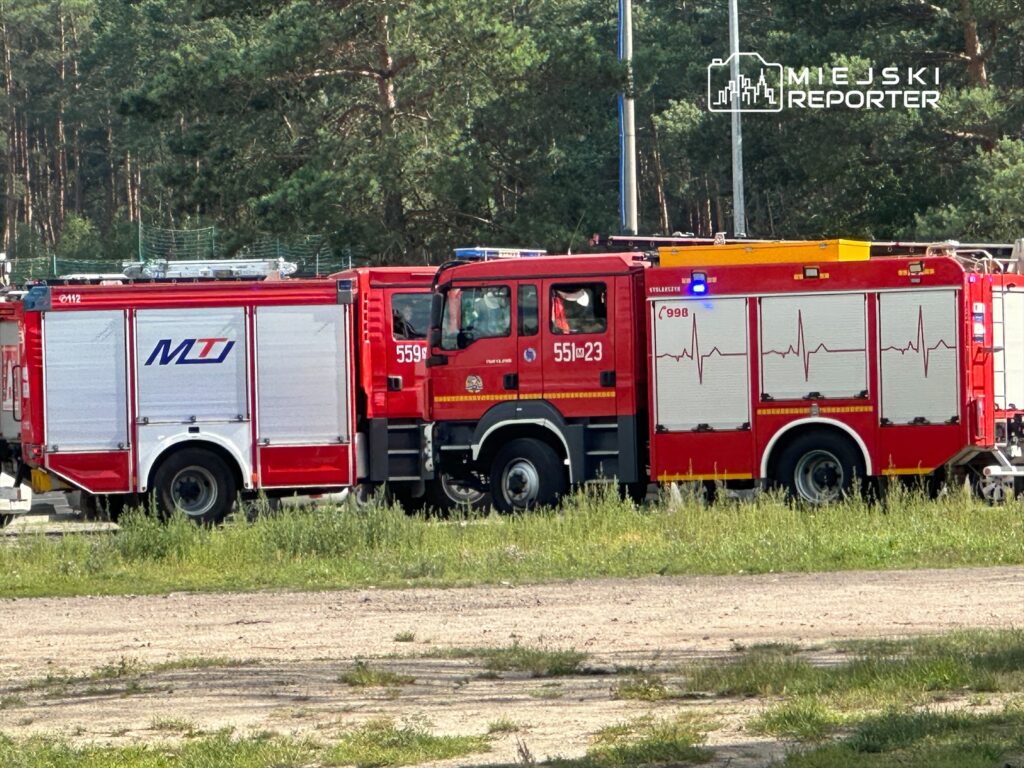 Dwa wozu strażackie zaparkowane na trawie, jeden z nich z napisem "MT" i niebieskim światłem.