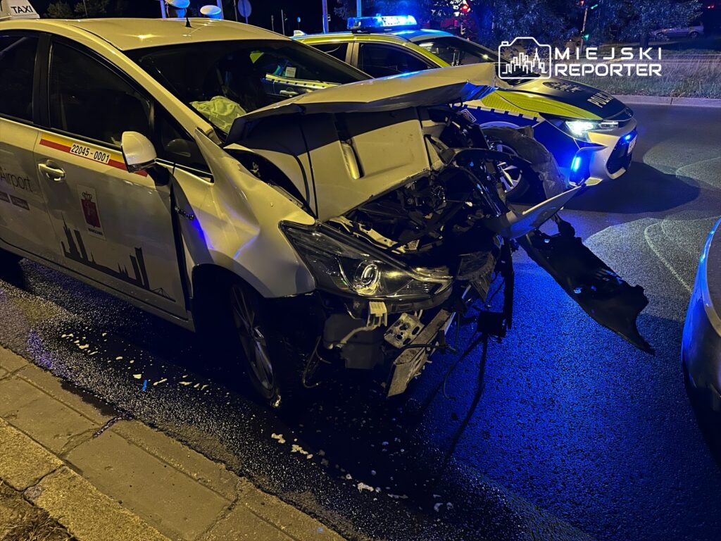 Zniszczony taksówka z uszkodzoną przednią częścią stoi na drodze, obok policyjny radiowóz z włączonymi sygnałami świetlnymi.
