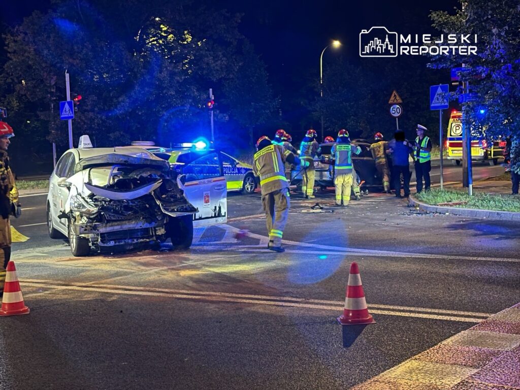 W nocy przy drodze, strażacy i policjanci pracują nad usunięciem skutków wypadku samochodowego, w tle widać uszkodzone pojazdy.