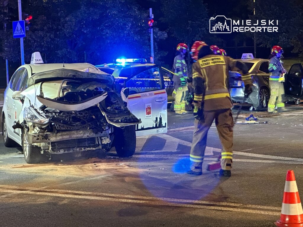 Zespół strażaków pracuje nad zabezpieczeniem miejsca wypadku drogowego z udziałem taksówki i osobowego samochodu.