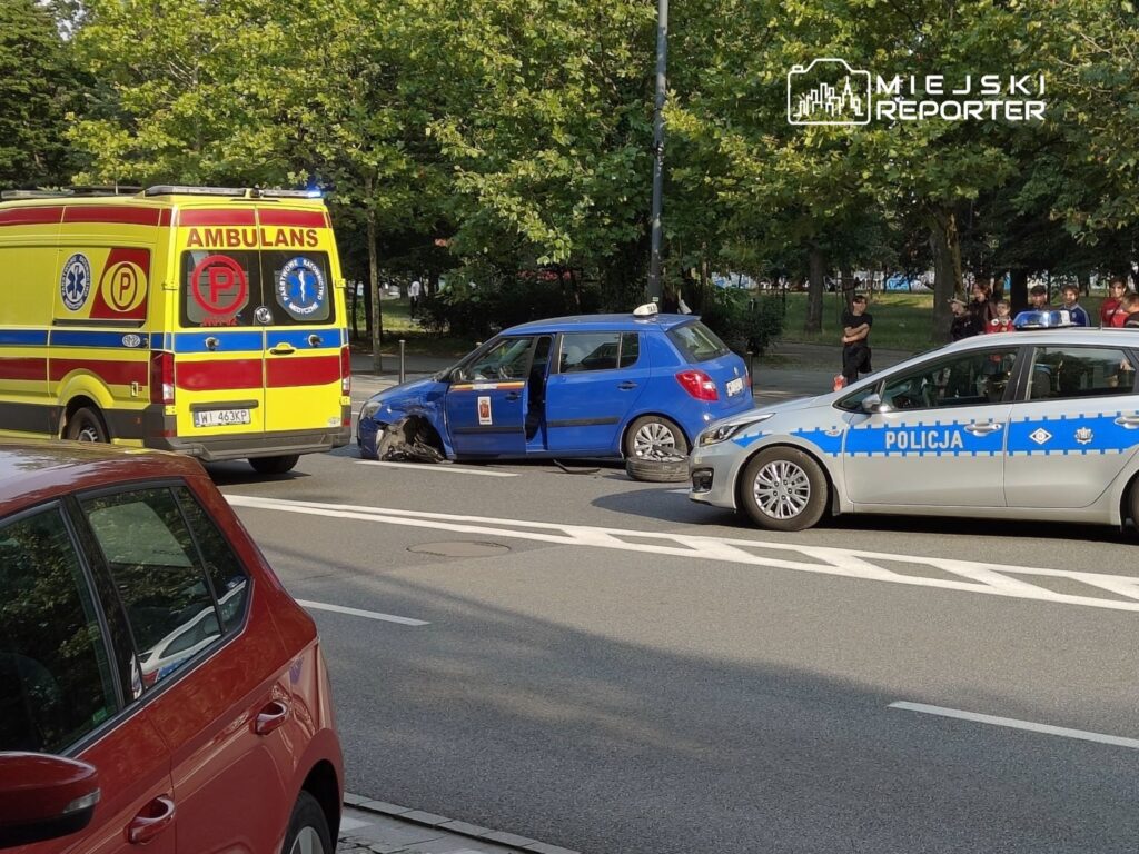 Ambulans i radiowóz policyjny stoją obok uszkodzonego niebieskiego samochodu na ulicy, w tle grupka ludzi.