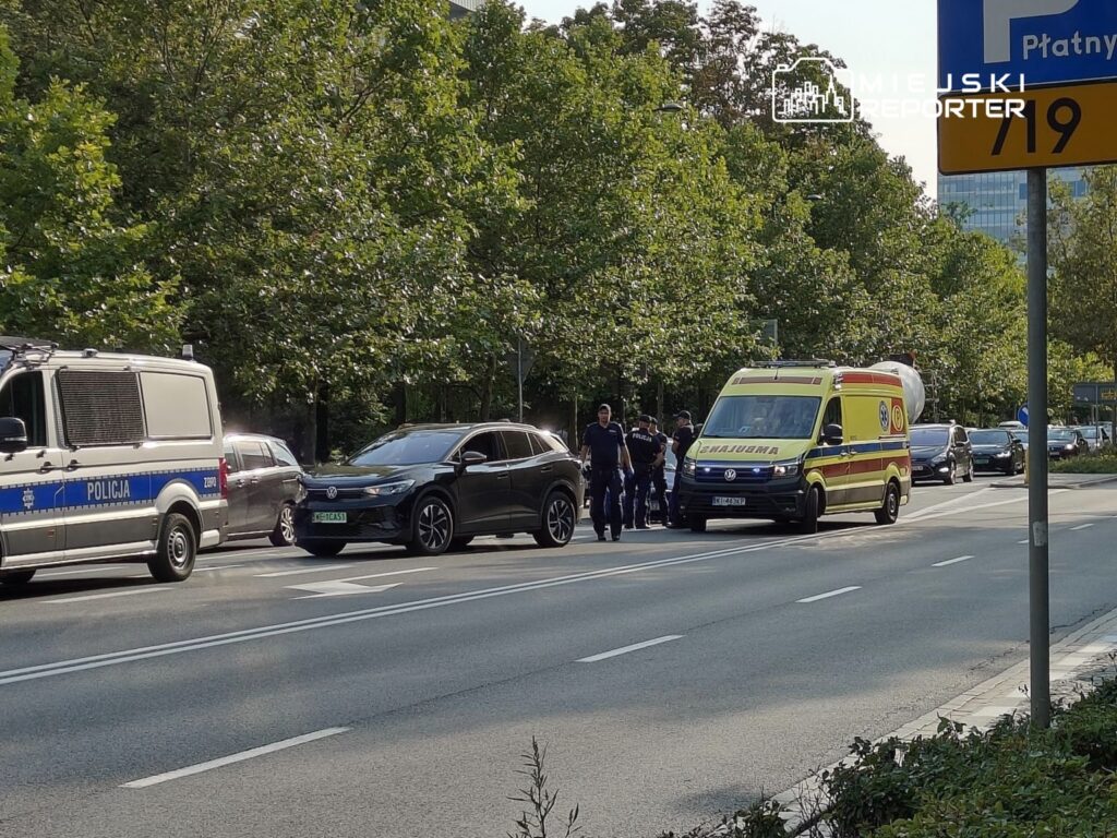 Policyjny radiowóz oraz karetka stoją na ruchliwej ulicy, obok grupy funkcjonariuszy rozmawiających z osobami w samochodach.