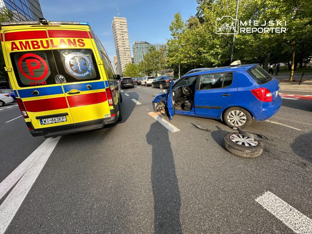 Niebieski samochód osobowy z otwartymi drzwiami stoi na jezdni obok karetki pogotowia, na drodze leży odcięte koło.