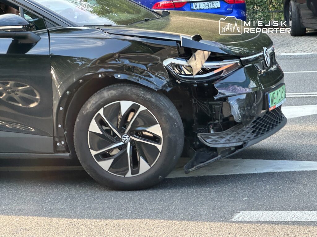Czarny samochód elektryczny z uszkodzoną przednią częścią stoi na ulicy, obok innych pojazdów.