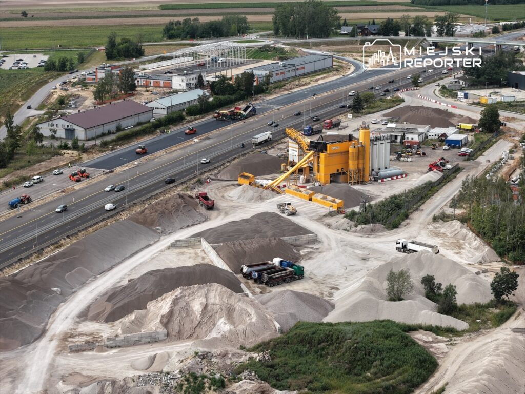 Koparki i ciężarówki transportujące materiały budowlane na placu budowy w pobliżu ruchliwej drogi.