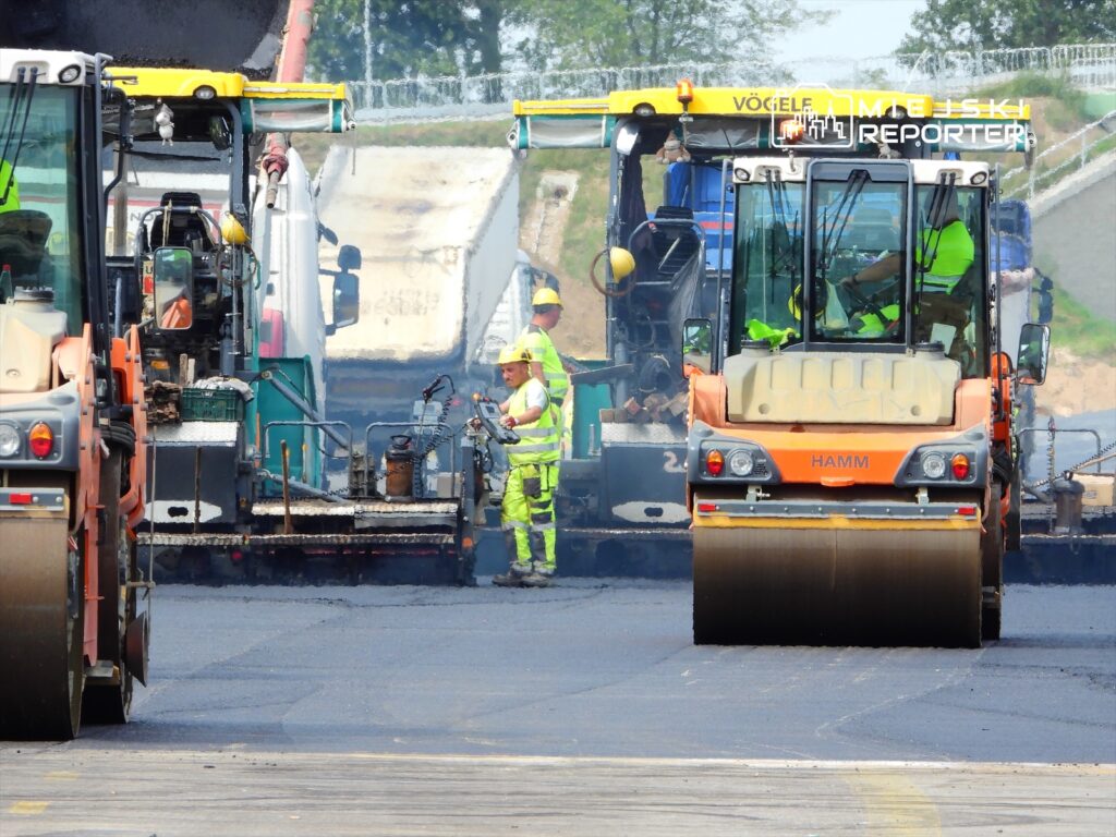 Grupa pracowników w odblaskowych kamizelkach nadzoruje prace drogowe z użyciem maszyn do asfaltowania.