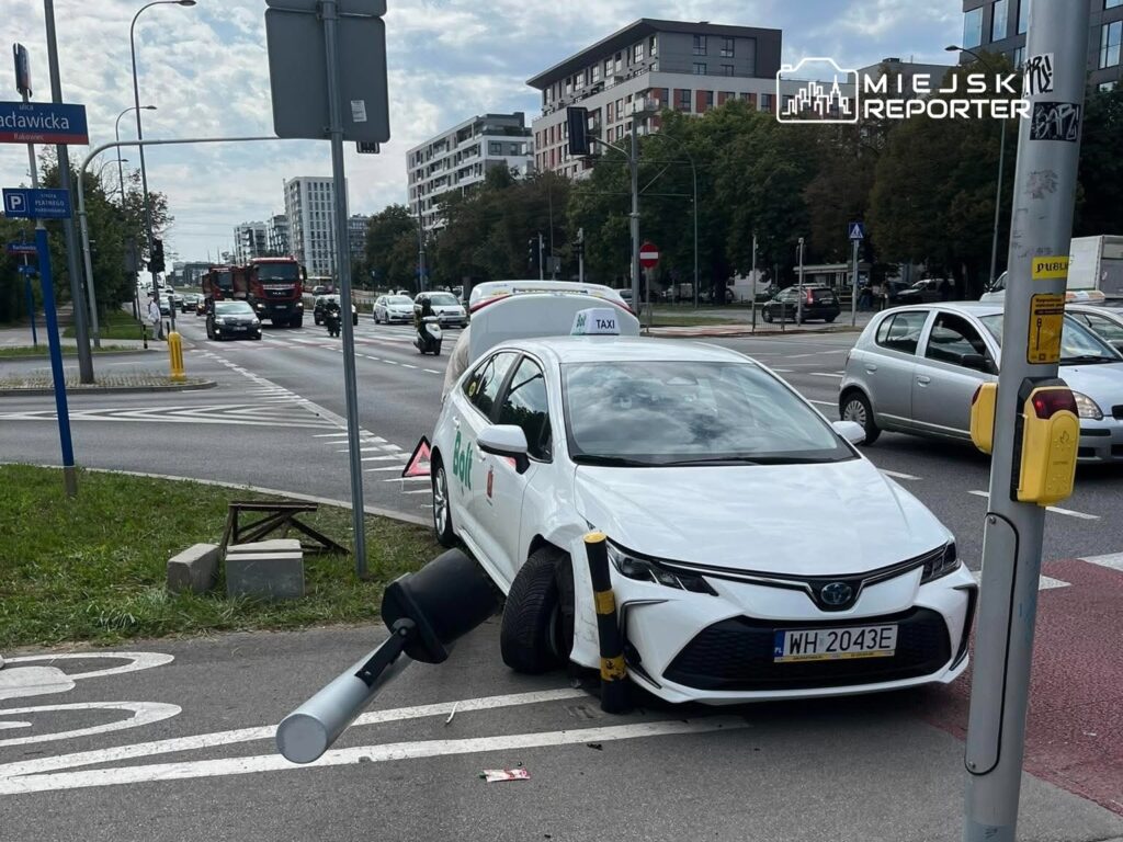 Biały samochód taxi z uszkodzoną przednią częścią stoi na chodniku, obok drogi z ruchem samochodowym.