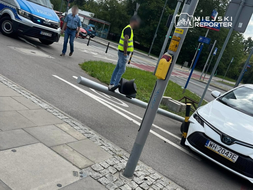 Policjant w niebieskim mundurze oraz mężczyzna w żółtej kamizelce sprawdzają przewrócony słup na ulicy.